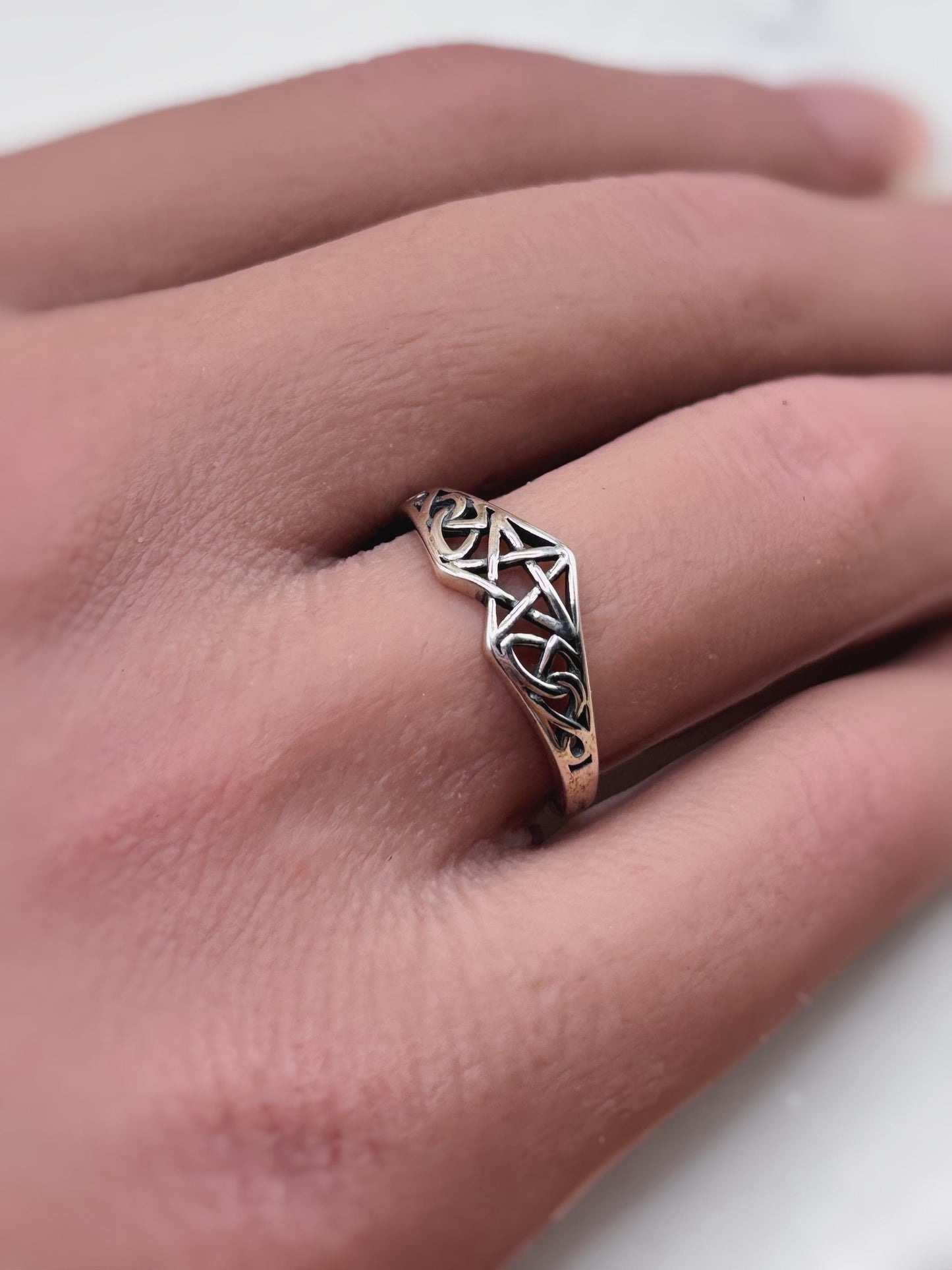 May include: A silver ring with an openwork design featuring a pentagram and Celtic knot patterns. The ring is worn on a finger, showcasing its intricate details and craftsmanship. The ring's design is geometric and symbolic.