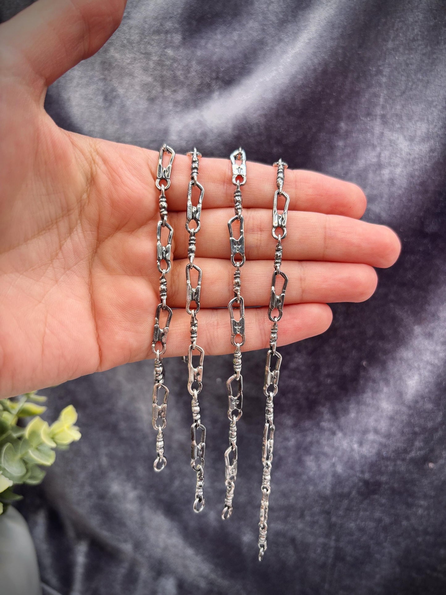 May include: Four silver-toned chain necklaces with a unique link design are displayed. The chains are held in a hand, showcasing their intricate details. The links have an irregular, textured appearance, adding to their handcrafted aesthetic. The background is a dark, textured fabric.