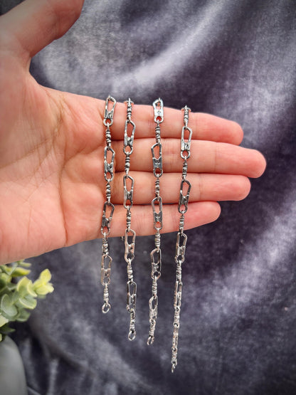 May include: Four silver-toned chain necklaces with a unique link design are displayed. The chains are held in a hand, showcasing their intricate details. The links have an irregular, textured appearance, adding to their handcrafted aesthetic. The background is a dark, textured fabric.