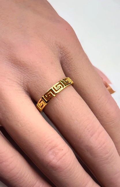 May include: A gold-colored ring featuring a repeating Greek key design. The ring is worn on a finger, showcasing its intricate pattern and shiny finish. The ring's design is a continuous, geometric pattern.