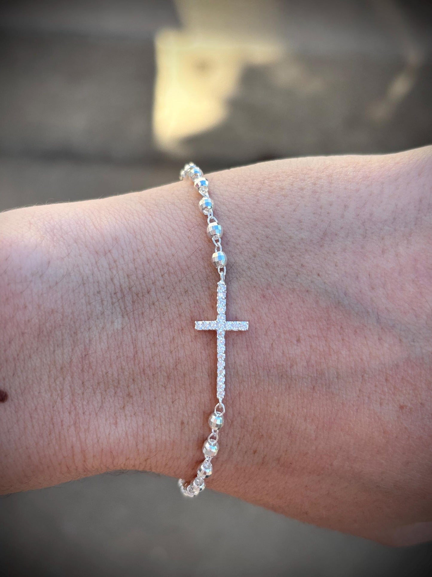 May include: A silver bracelet featuring a cross pendant encrusted with small, sparkling stones. The bracelet is composed of small, round beads and is designed to be worn on the wrist. The cross is the focal point of the bracelet.