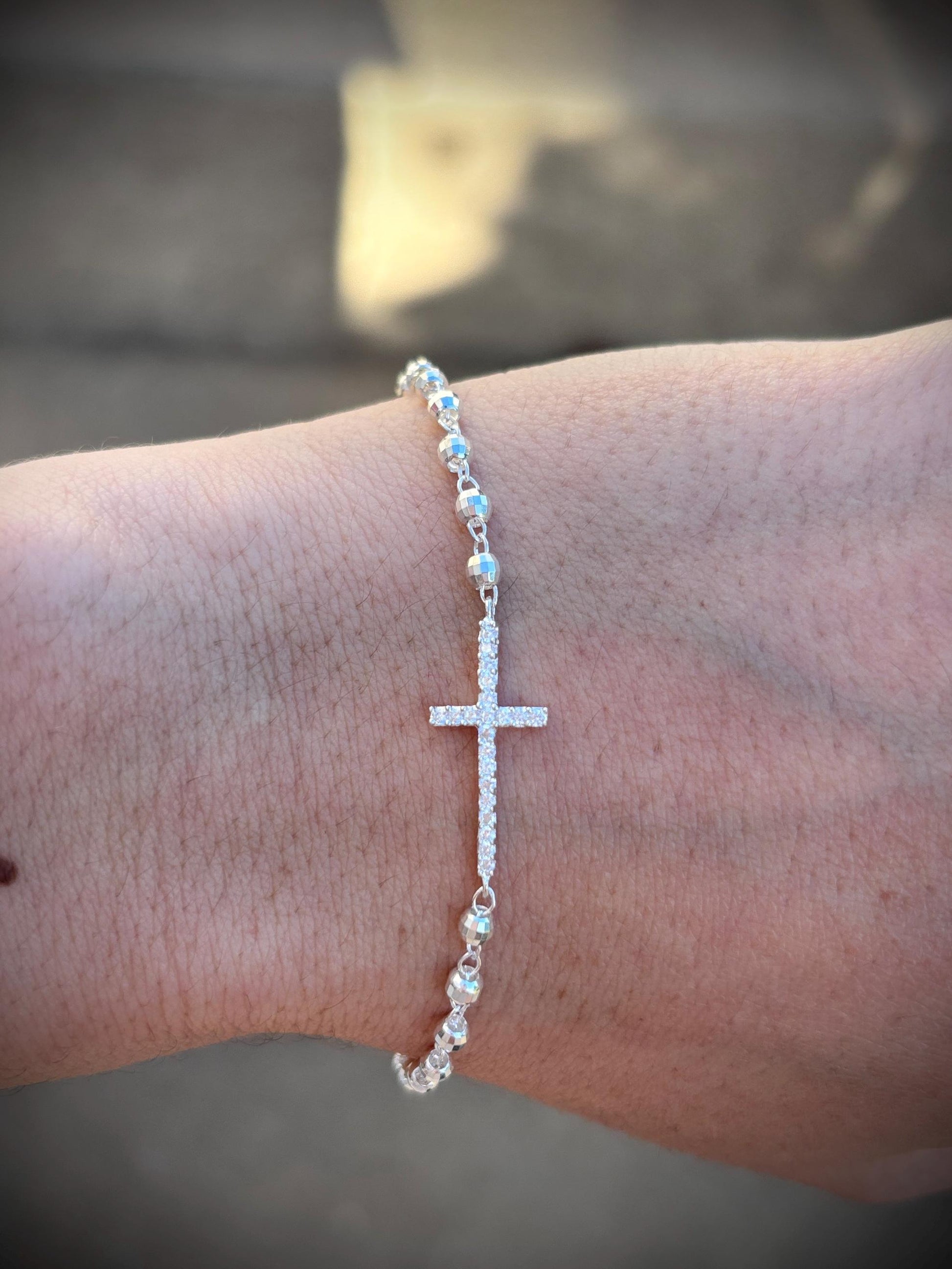 May include: A silver bracelet featuring a cross pendant encrusted with small, sparkling stones. The bracelet is composed of small, round beads and is designed to be worn on the wrist. The cross is the focal point of the bracelet.