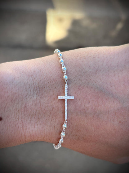 May include: A silver bracelet featuring a cross pendant encrusted with small, sparkling stones. The bracelet is composed of small, round beads and is designed to be worn on the wrist. The cross is the focal point of the bracelet.