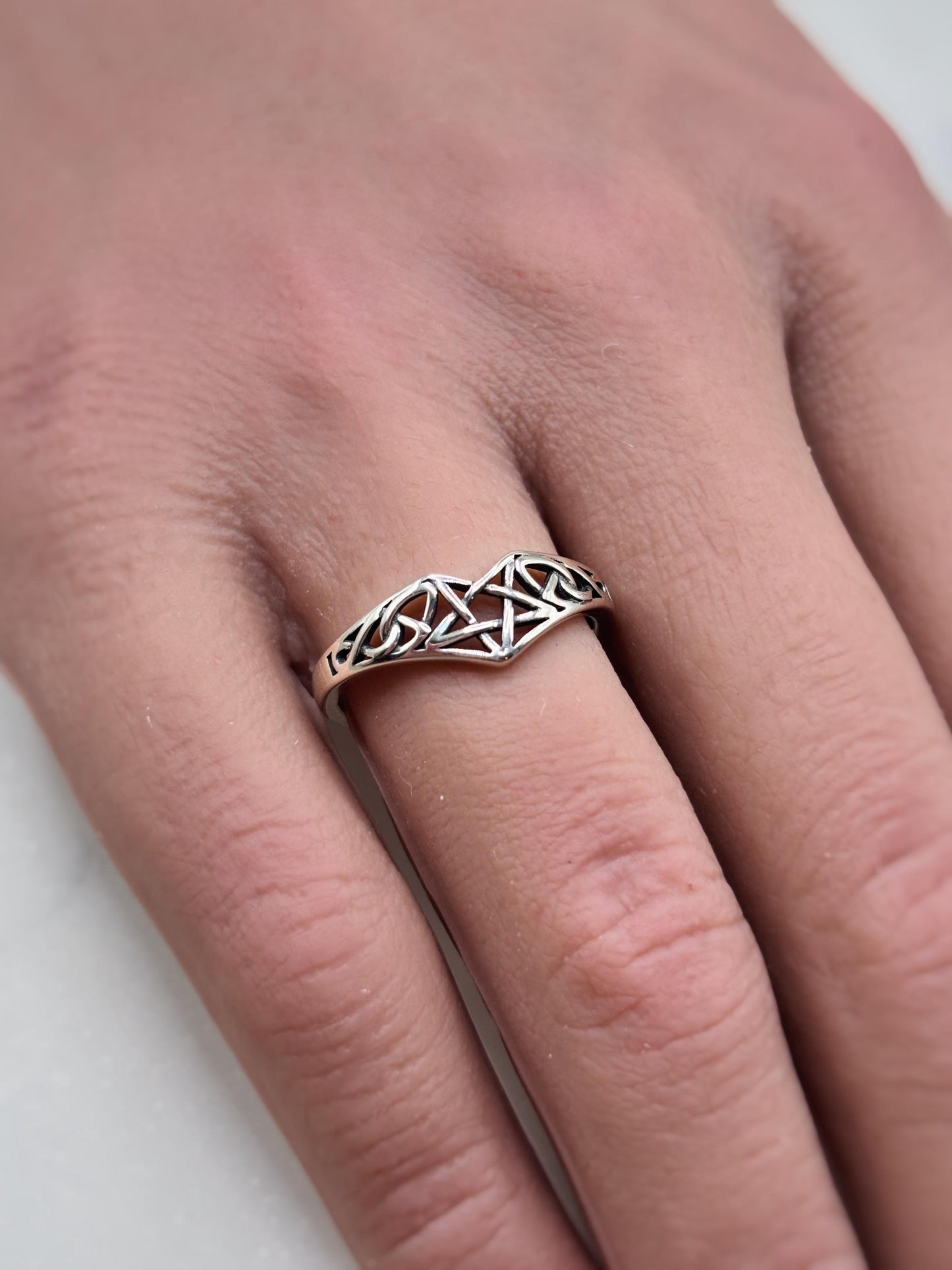 May include: A silver-colored ring with a Celtic knot design. The ring features an intricate pattern of interwoven lines forming a decorative band. The ring is worn on a finger, showcasing the detailed craftsmanship and the ring's elegant style.