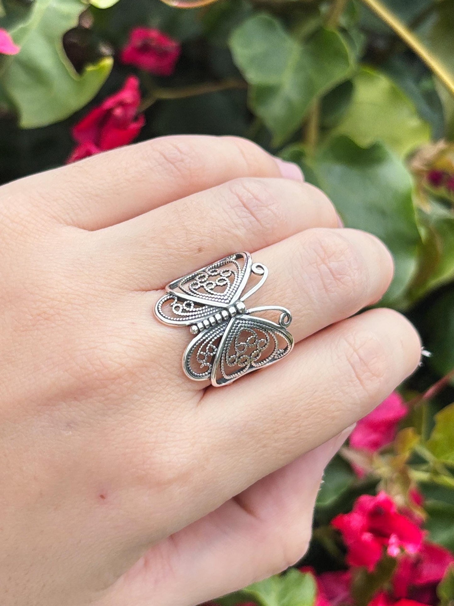 May include: A silver butterfly ring with intricate filigree detailing. The ring features a delicate openwork design, showcasing the butterfly's wings and body. The ring is worn on a finger, with a blurred background of green foliage and pink flowers.
