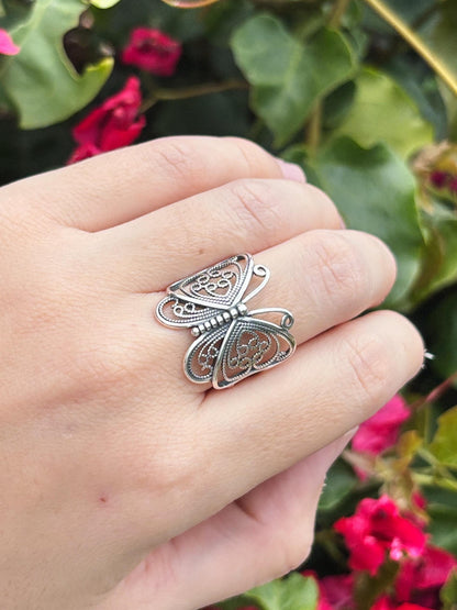 May include: A silver butterfly ring with intricate filigree detailing. The ring features a delicate openwork design, showcasing the butterfly's wings and body. The ring is worn on a finger, with a blurred background of green foliage and pink flowers.