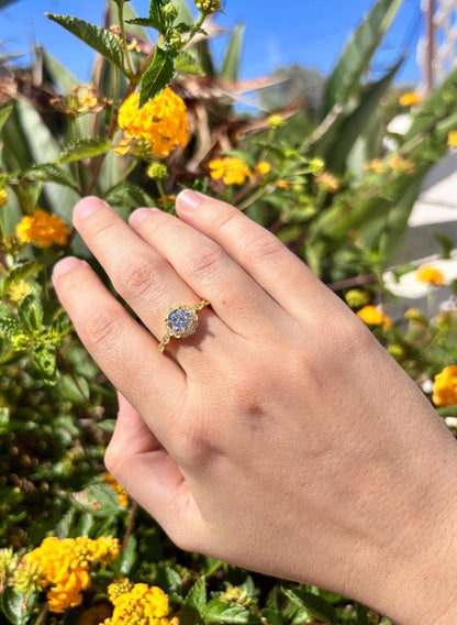 May include: A gold ring with a round, light blue gemstone set in a decorative bezel. The ring is on a hand, with yellow flowers and green foliage in the background. The ring's band has a twisted rope design.