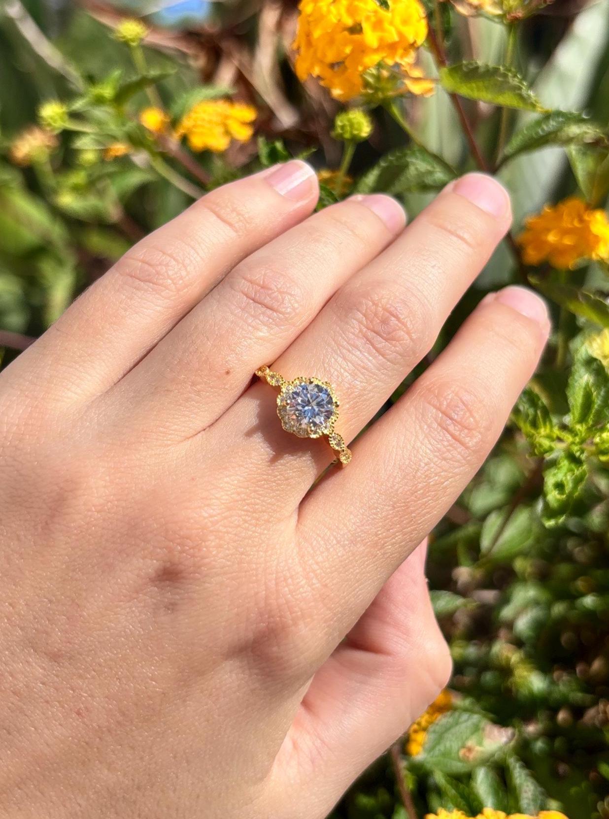 May include: A gold ring with a large, round, clear gemstone at the center, surrounded by smaller clear stones. The ring has a detailed, ornate design. The background features green leaves and yellow flowers.