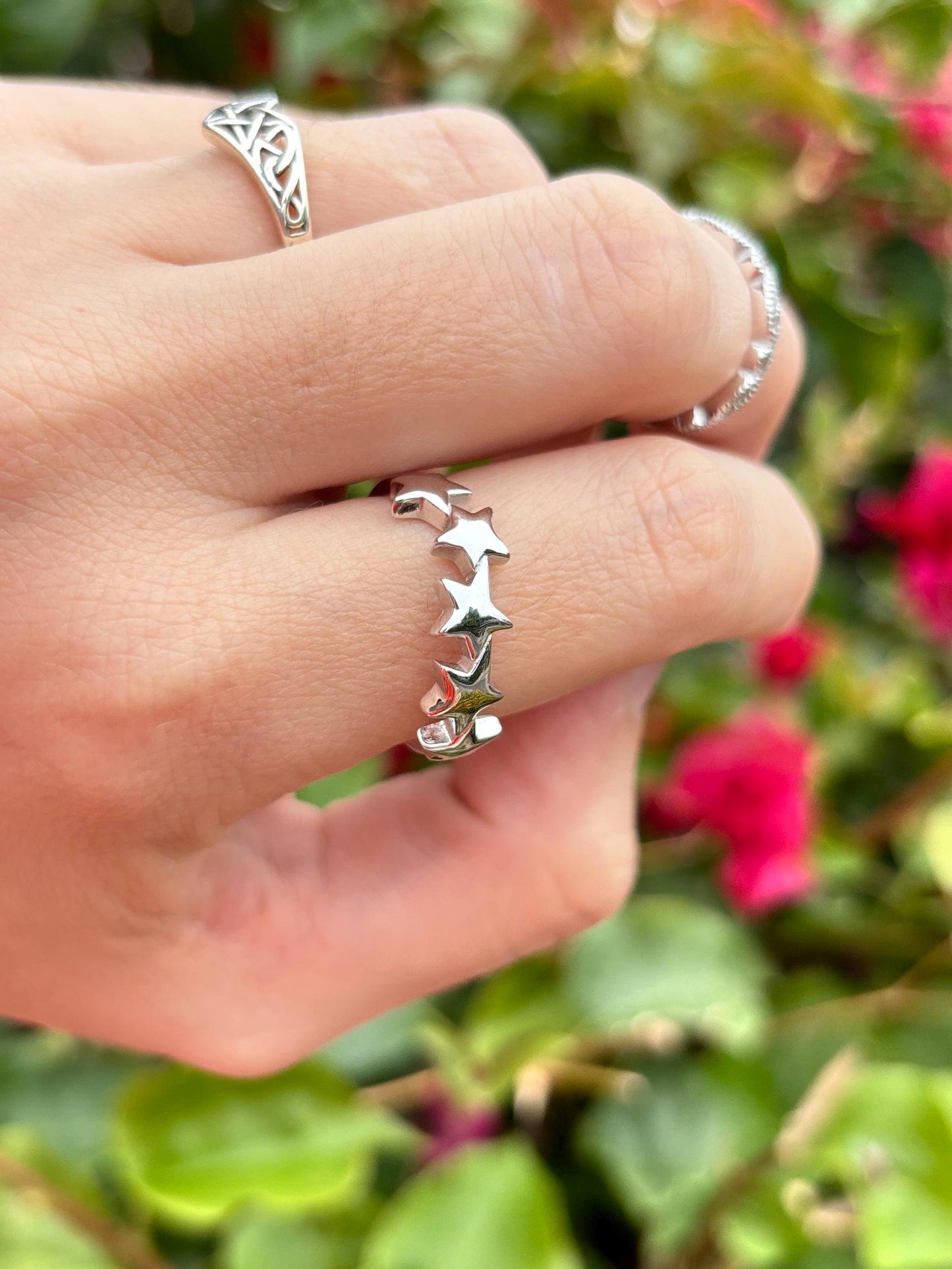 May include: A silver ring with a row of five star-shaped cutouts. The ring is on a finger, with two other silver rings visible in the background. The background is blurred with green foliage and pink flowers.