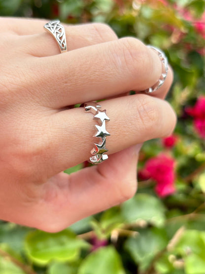 May include: A silver ring with a row of five star-shaped cutouts. The ring is on a finger, with two other silver rings visible in the background. The background is blurred with green foliage and pink flowers.