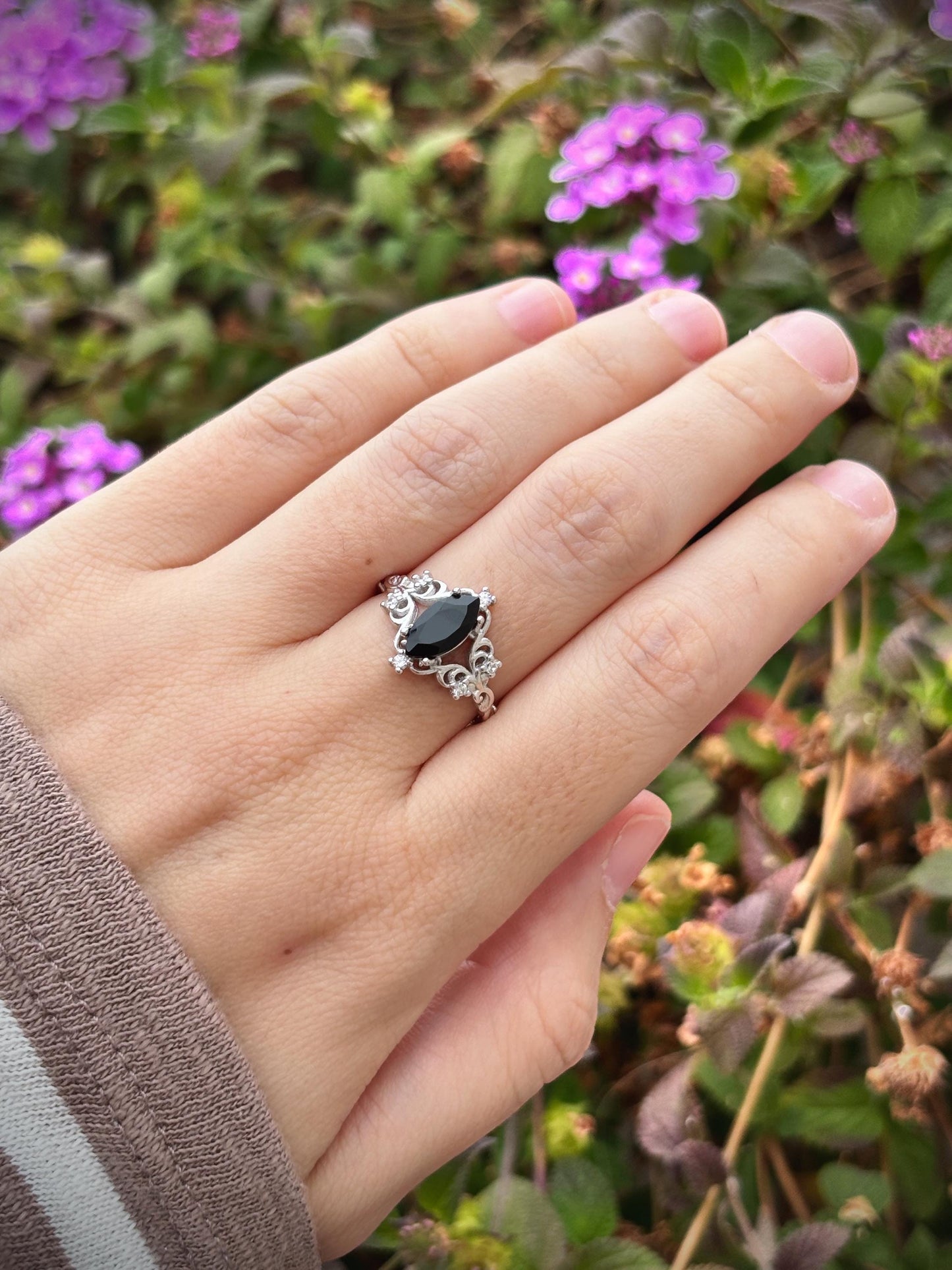 May include: A silver ring with a black marquise-cut stone, surrounded by small white stones and ornate detailing. The ring is set against a blurred background of green foliage and purple flowers.
