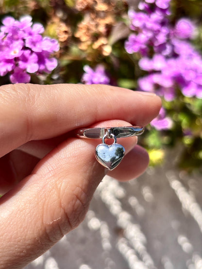 May include: A silver ring with a small, dangling heart charm. The ring is held up against a blurred background of purple flowers and green foliage, creating a soft, natural setting. The heart charm is polished and reflective.