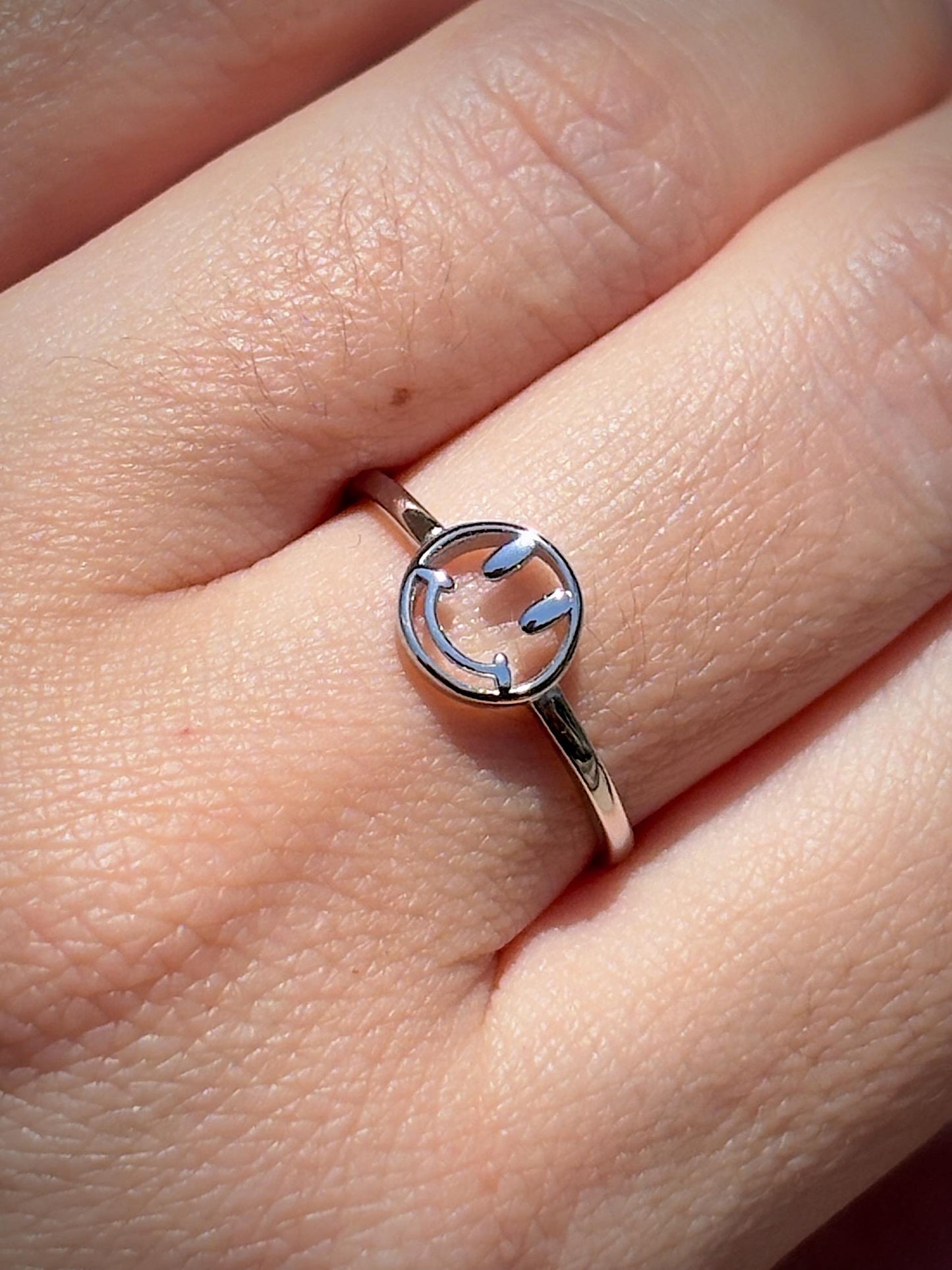 May include: A silver ring featuring a smiley face design. The ring has a thin band and a circular face with cut-out eyes and a curved mouth. The ring is worn on a finger.