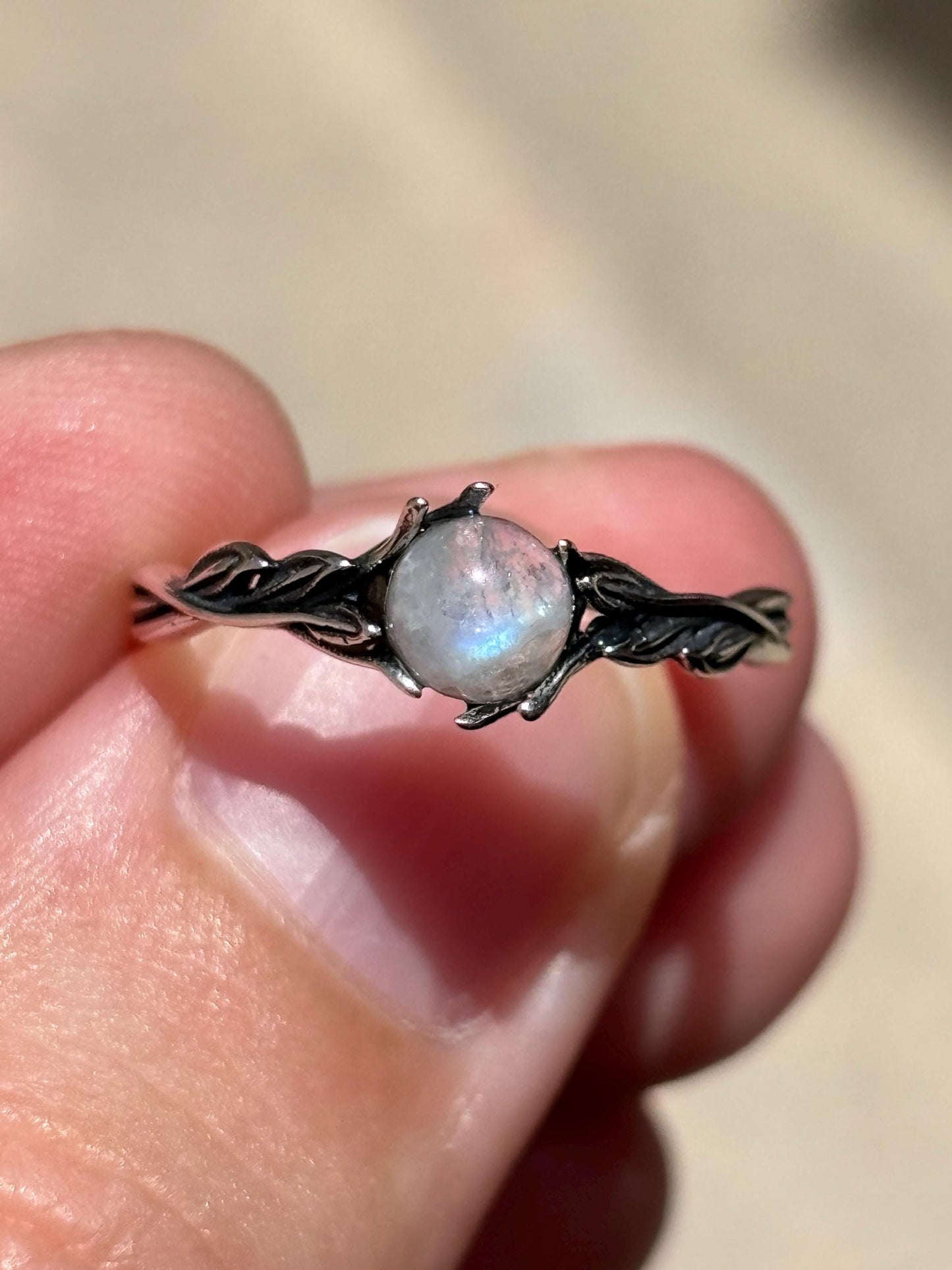 May include: A silver ring featuring a round, iridescent gemstone. The ring's band is designed with a delicate, intertwined leaf pattern. The gemstone is the focal point, showcasing a play of colors.