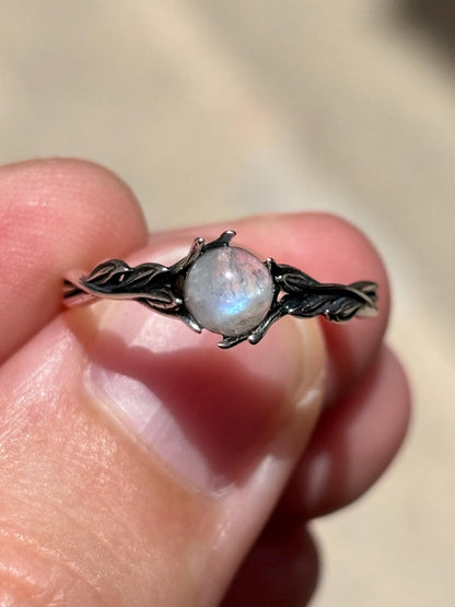 May include: A silver ring featuring a round, iridescent gemstone. The ring's band is designed with a delicate, intertwined leaf pattern. The gemstone is the focal point, showcasing a play of colors.