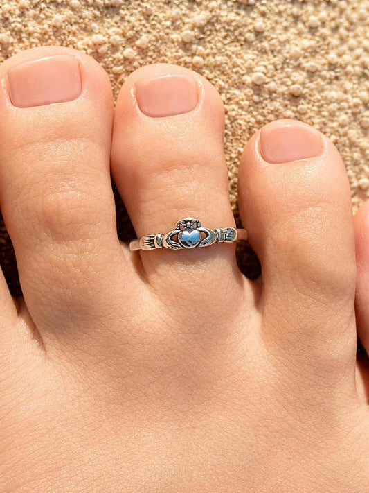 May include: A silver toe ring featuring a Claddagh design with a heart-shaped center. The ring is worn on a foot with manicured nails, set against a sandy background. The Claddagh ring symbolizes love, loyalty, and friendship.