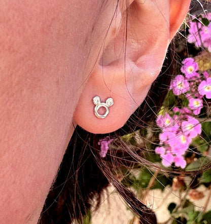 May include: A silver stud earring in the shape of a mouse head is shown. The earring has a circular center with two smaller circles above it, forming the mouse's ears. The earring is on an ear, with pink flowers in the background.