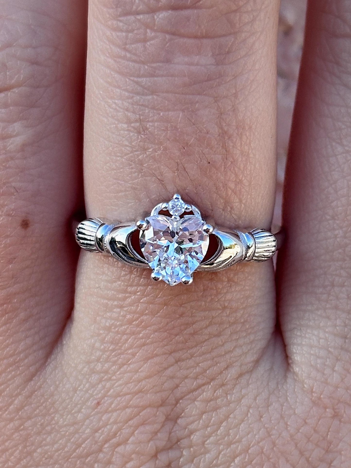 May include: A silver-toned Claddagh ring featuring a heart-shaped, clear gemstone. The ring's design includes two hands clasping a heart, topped with a crown. The ring is worn on a finger.