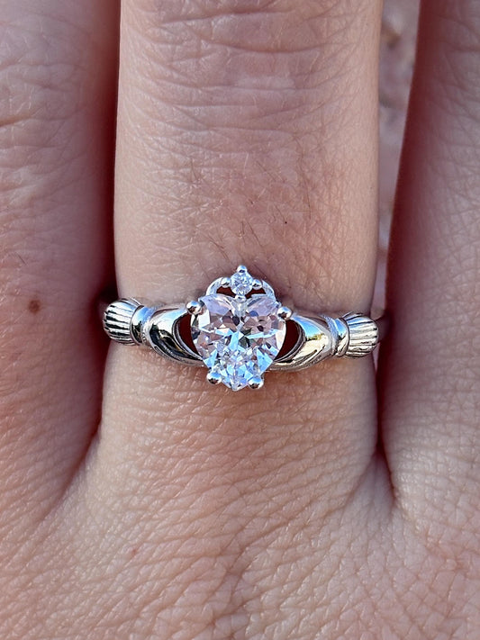 May include: A silver-toned Claddagh ring featuring a heart-shaped, clear gemstone. The ring's design includes two hands clasping a heart, topped with a crown. The ring is worn on a finger.