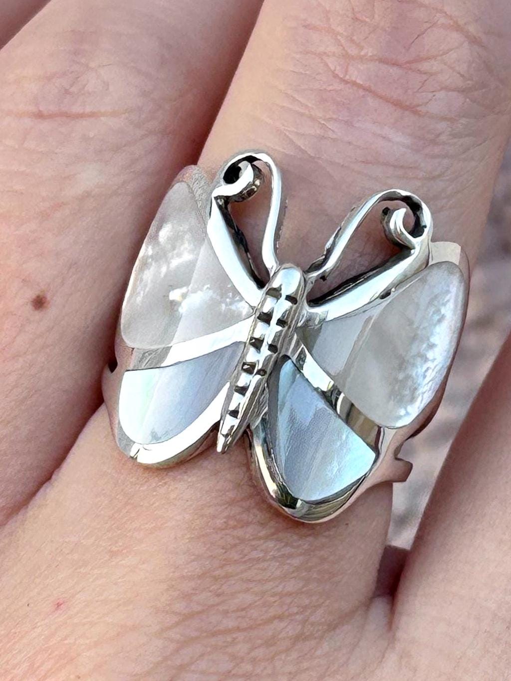 May include: A silver ring shaped like a butterfly with iridescent white mother-of-pearl inlays on the wings. The metal body of the butterfly features a segmented design, and the upper wing sections have curved, open-work silver detailing.