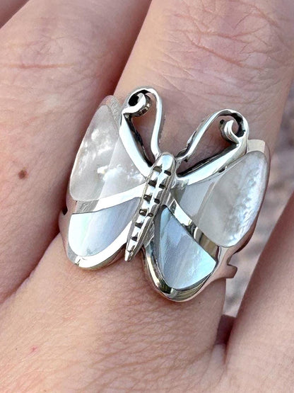 May include: A silver ring shaped like a butterfly with iridescent white mother-of-pearl inlays on the wings. The metal body of the butterfly features a segmented design, and the upper wing sections have curved, open-work silver detailing.