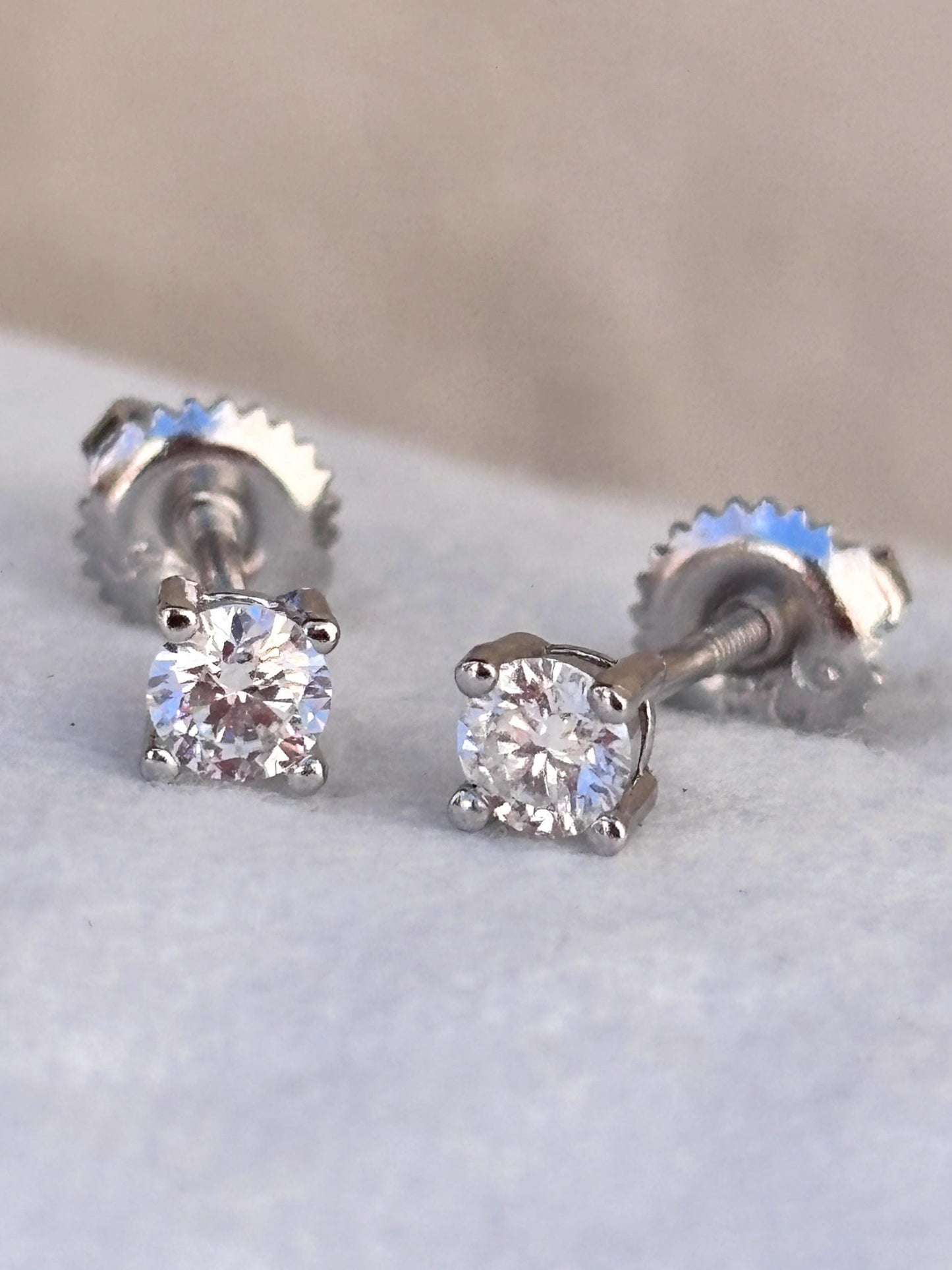 May include: A pair of sparkling diamond stud earrings. Each earring features a round, brilliant-cut diamond set in a four-prong setting. The posts have a screw-back closure. The earrings are set against a soft, neutral background.