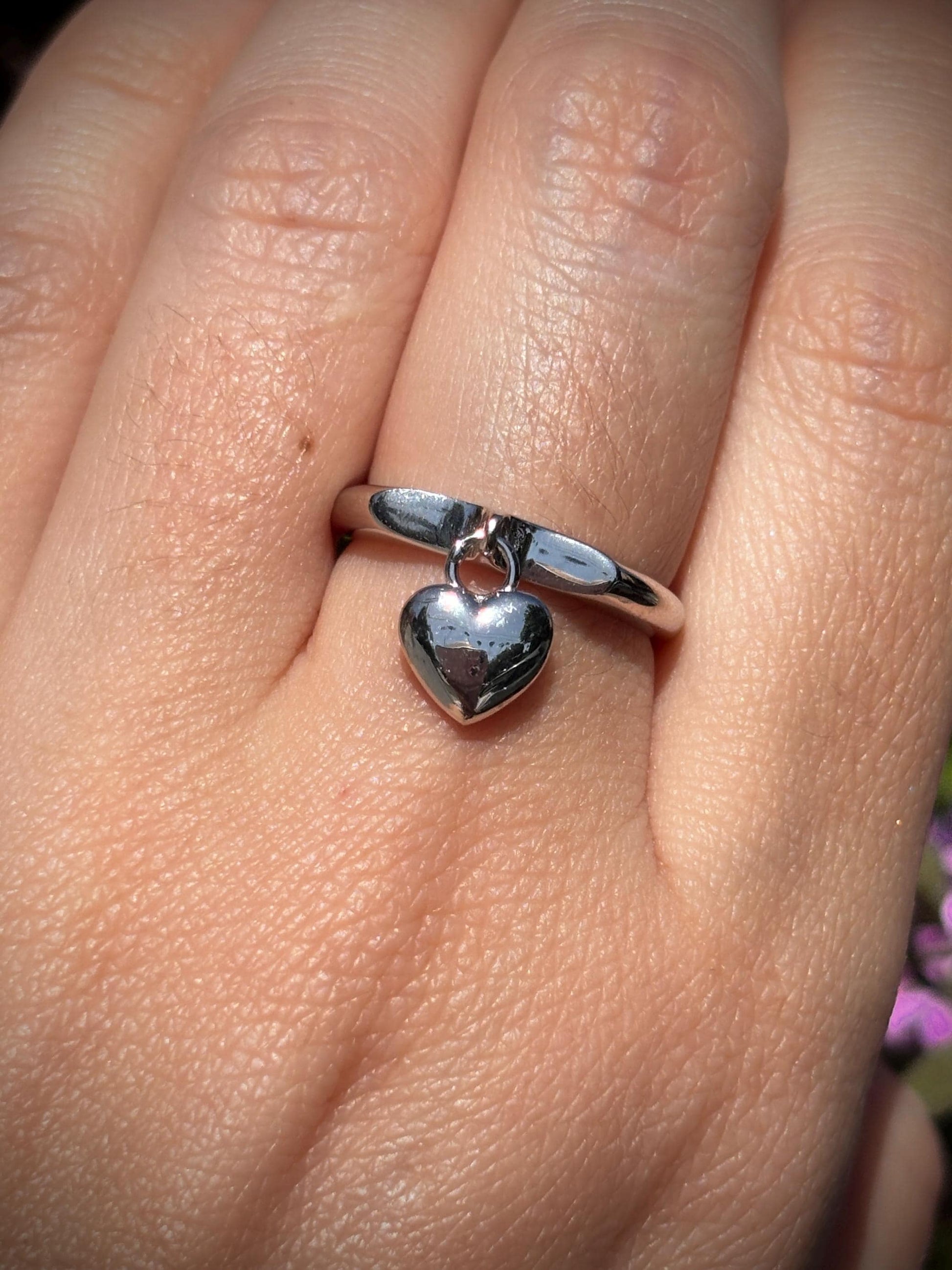 May include: A silver ring with a dangling heart charm. The heart is polished and reflects light. The ring is simple and elegant, suitable for everyday wear or special occasions.
