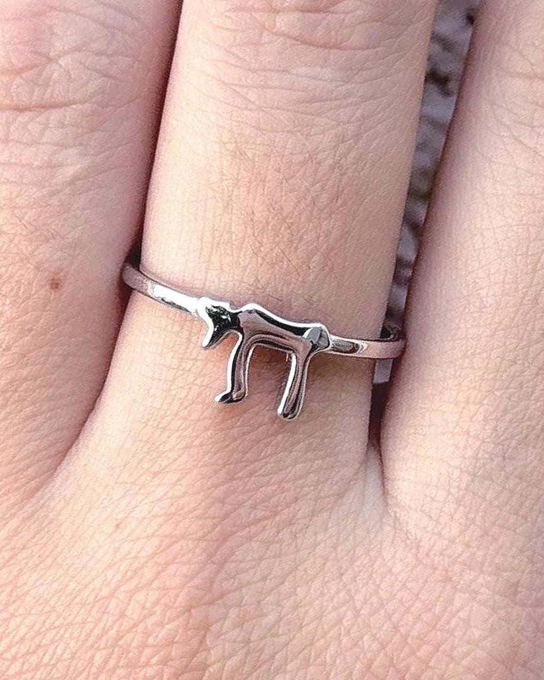 May include: A silver-colored ring featuring a stylized Hebrew letter. The ring is thin and delicate, with the letter prominently displayed. The ring is worn on a finger, showcasing its simple yet meaningful design.