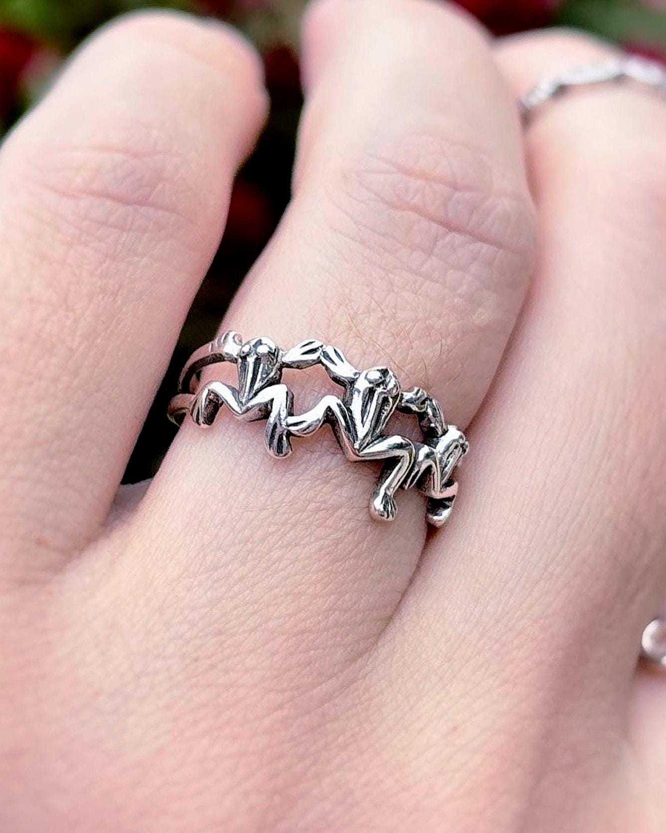 May include: A silver ring with a repeating frog design is worn on a finger. Another ring with an infinity symbol design is also visible. The background features green foliage and vibrant pink flowers.