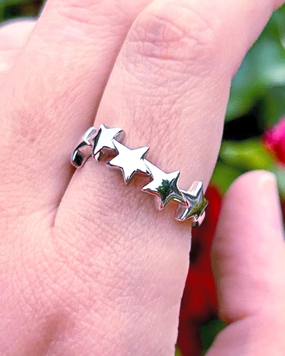 May include: A silver ring featuring a row of three star-shaped designs. The ring is worn on a finger, with other rings visible in the background. The image is taken in natural light, with a blurred background of green foliage and red flowers.