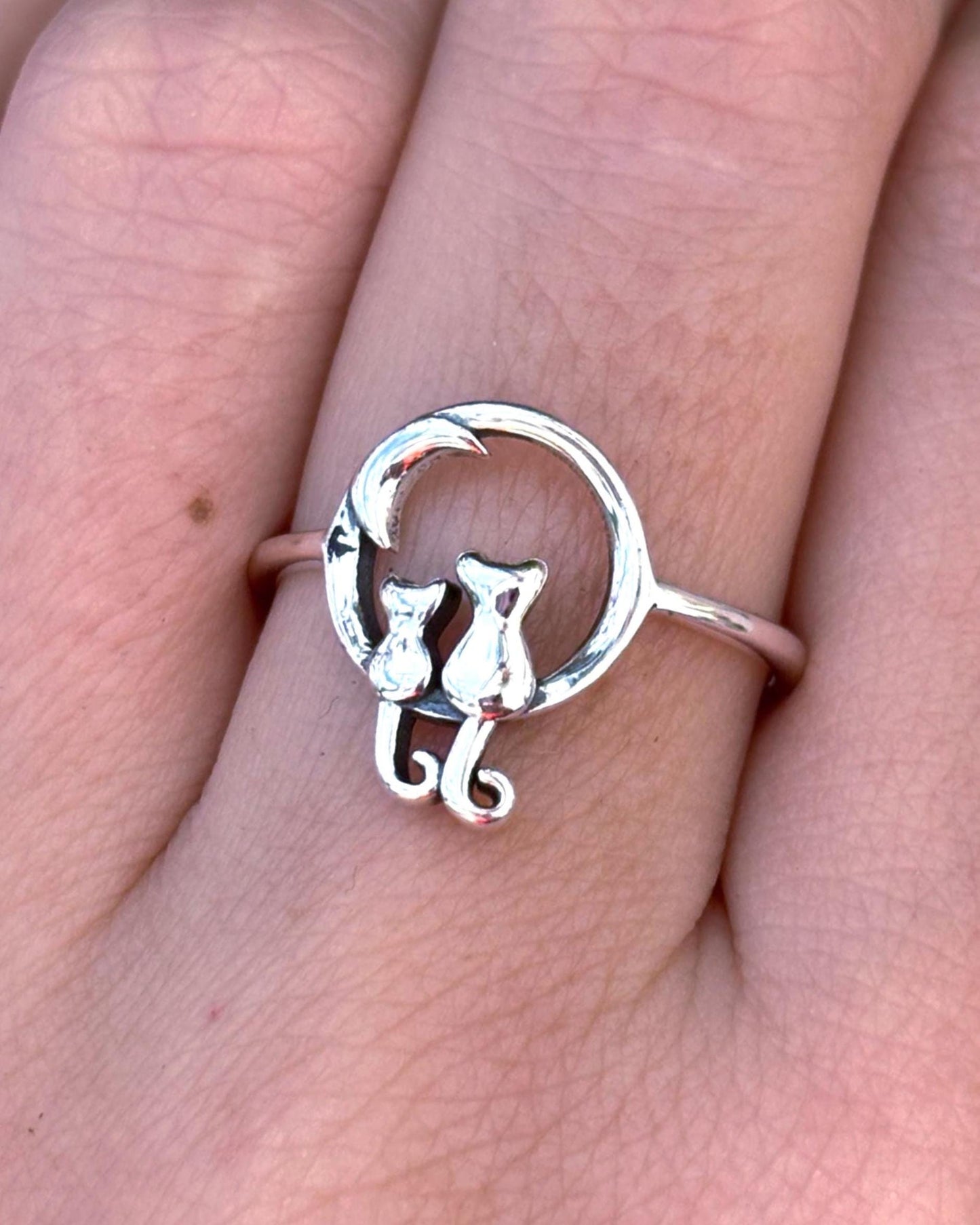 May include: A silver ring featuring a circular design with a crescent moon and two cat silhouettes sitting side by side. The cats are positioned at the base of the moon, with their tails curved downward, creating a continuous metallic band.